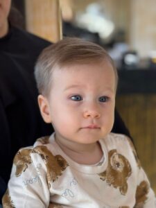 Imagine de aproape cu un bebelus cu par blond fin, tuns ingrijit si dat intr-o parte, cu contur curat pe laterale. Parul este uniform si bine aranjat, iar bebelusul poarta o hainuta alba cu imprimeu. In fundal se observa spatiul salonului Sia's Beauty din Lunca Cetatuii. Imaginea surprinde rezultatul unei tunsori delicate realizate la frizerie copii Iasi, cu atentie si grija pentru cei mai mici clienti.