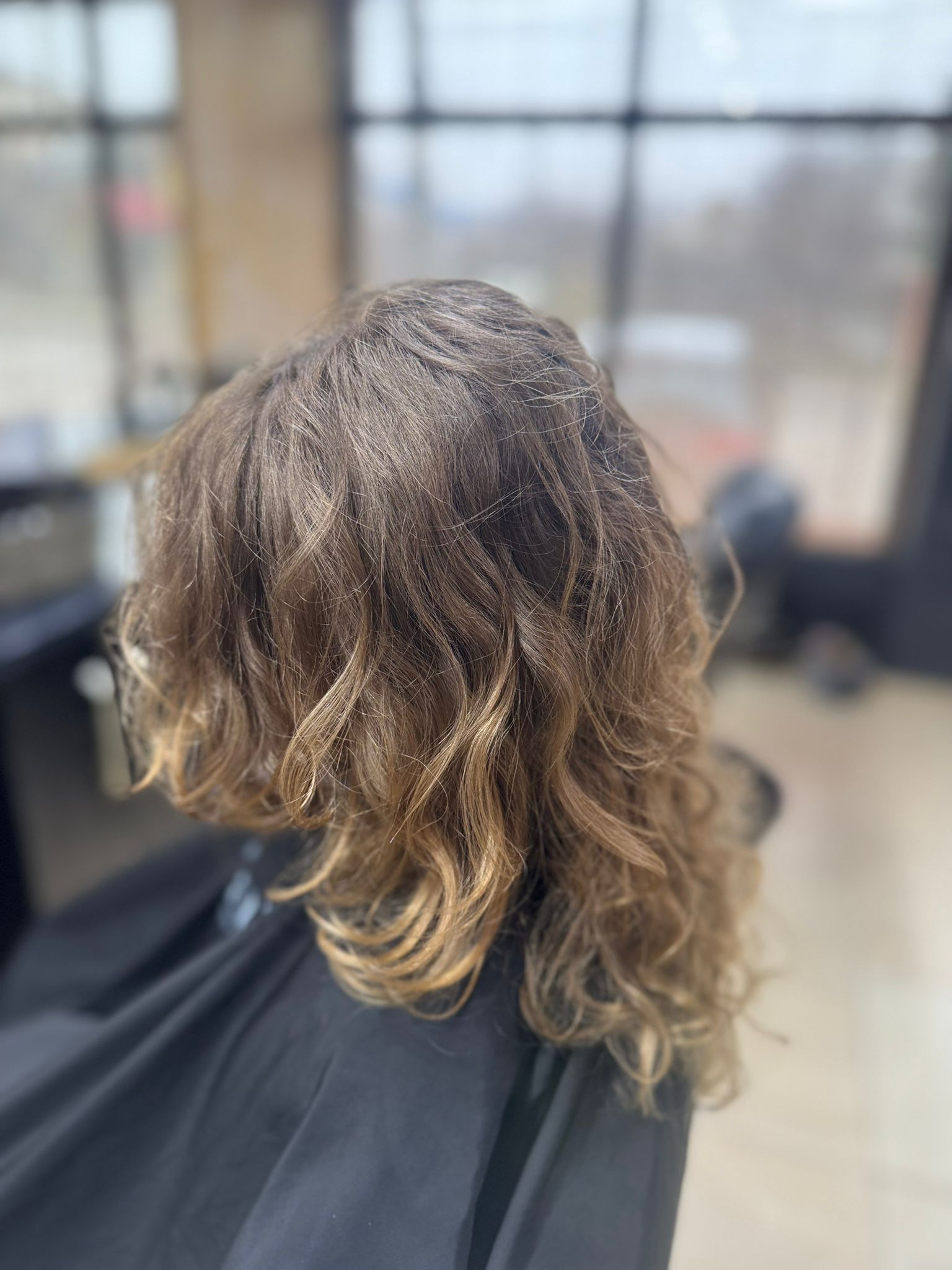 Vedere din spate a unei tunsori pentru femei cu bucle lejere si naturale, par castaniu deschis cu reflexe blonde aurii la vârfuri si volum, realizata la Sia's Beauty Iasi, Lunca Cetatuii.