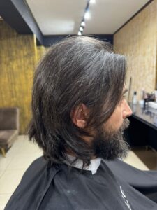 Imagine in profil a unui barbat cu par lung inchis la culoare, umed, pregatit pentru tuns, si barba densa. Clientul sta in scaunul salonului cu un halat negru de protectie. In fundal se observa interiorul salonului Sia's Beauty cu pereti decorativi, canapea si statie de lucru cu produse specifice unui barbershop Iasi din Lunca Cetatuii. Imaginea surprinde momentul dinaintea tunsoarei.