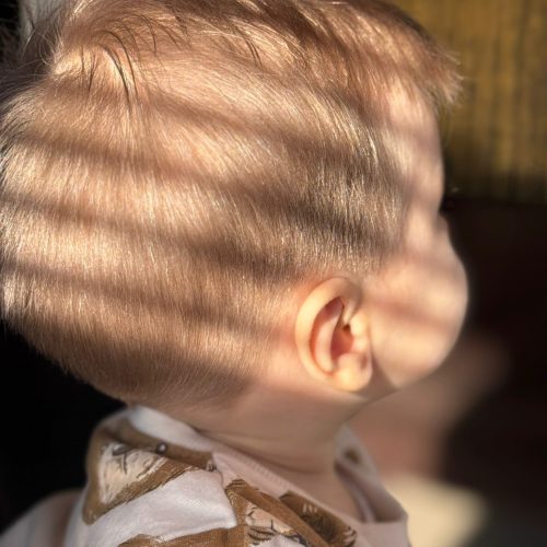 Imagine in profil a unui bebelus cu par blond fin, tuns ingrijit si uniform, cu contur curat in jurul urechii. Parul este dat natural spre spate, cu o trecere blanda pe laterale. Bebelusul poarta o hainuta cu imprimeu colorat. Lumina naturala evidentiaza fineta parului si ingrijirea atenta cu care a fost realizata tunsoarea. Imaginea surprinde una dintre cele mai speciale vizite la frizerie copii Iasi, la Sia's Beauty din Lunca Cetatuii.