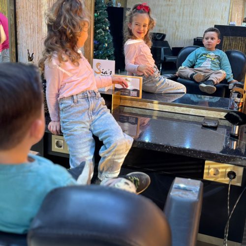 Doi copii se joaca si interactioneaza relaxat in interiorul salonului Sia's Beauty, in asteptarea randului la tuns. O fetita cu par cret si accesorii rosii in par sta pe tejgheaua salonului, iar un baietel sta comod in scaunul de barbershop. In fundal se observa bradul de Craciun, logo-ul Sia's Beauty Center si interiorul caracteristic al salonului din Lunca Cetatuii. Imaginea surprinde atmosfera calda si primitoare a salonului de frizerie copii Iasi.