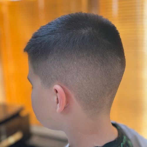 Imagine din spate si lateral a unui baiat cu o tunsoare tip skin fade executata foarte curat, cu degradeu fin care porneste aproape de la piele pe laterale si ceafa si se continua treptat spre parul scurt de deasupra. Conturul cefei este precis si uniform. Baiatul poarta un halat alb de protectie. Fundalul cald, portocaliu, apartine salonului Sia's Beauty din Lunca Cetatuii, unde frizerie copii Iasi se realizeaza cu aceeasi atentie ca pentru clientii adulti.