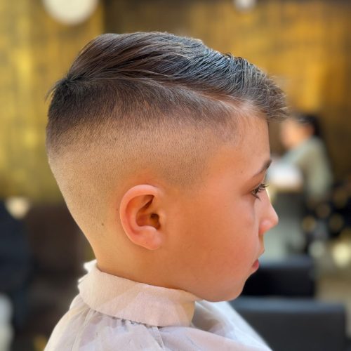 Imagine in profil a unui baiat cu o tunsoare moderna tip slick back cu high fade, cu degradeu ridicat si curat pe laterale si ceafa, si par mai lung deasupra dat spre spate si coafat ingrijit. Linia de separatie dintre zonele trase la masina si parul de deasupra este precisa si bine conturata. Baiatul poarta un halat alb de protectie. In fundal se observa peretele auriu decorativ si scaunele negre specifice salonului Sia's Beauty din Lunca Cetatuii. Tunsoarea a fost realizata la frizerie copii Iasi.