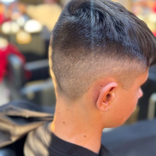 Imagine din spate si lateral a unui baiat cu o tunsoare moderna tip fade, cu degradeu curat pe laterale si ceafa, si par mai lung deasupra, dat spre spate cu volum. Trecerea dintre zonele trase la masina si parul de deasupra este precisa si bine executata. Baiatul poarta un halat negru de protectie. In fundal se observa oglinzile si luminile rotunde specifice salonului Sia's Beauty. Tunsoarea a fost realizata la frizerie copii Iasi, Sia's Beauty din Lunca Cetatuii.