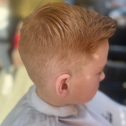 Imagine din spate si lateral a unui baiat mic cu par roscat natural, tuns ingrijit cu un degradeu curat pe laterale si ceafa si par mai lung deasupra, dat spre spate si coafat cu volum. Trecerea dintre zonele trase la masina si parul de deasupra este fina si precisa. Baiatul poarta un halat alb de protectie. Imaginea surprinde rezultatul final al unei tunsori realizate la frizerie copii Iasi, la Sia's Beauty din Lunca Cetatuii.