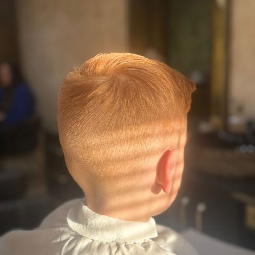 Imagine din spate a unui baiat mic cu par roscat natural, tuns ingrijit, cu degradeu fin pe laterale si ceafa si par mai lung deasupra, dat usor spre fata. Conturul cefei este curat si bine definit. Baiatul poarta un halat alb de protectie. Lumina naturala evidentiaza culoarea naturala a parului si calitatea tunsoarei realizate la frizerie copii Iasi, la Sia's Beauty din Lunca Cetatuii.
