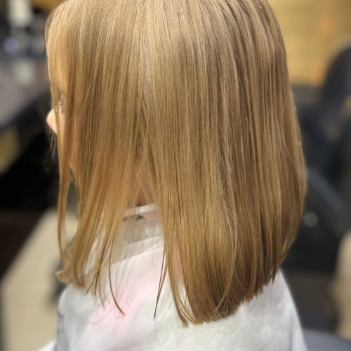 Imagine din spate a unei cliente cu par blond auriu, drept si stralucitor, tuns in forma de bob cu linie dreapta la baza. Parul este uniform, bine ingrijit si coborat pana la nivelul umerilor, cu o trecere naturala de culoare de la radacina spre varf. Clienta poarta un halat alb de protectie. In fundal se observa spatiul luminos al salonului Sia's Beauty din Lunca Cetatuii. Imaginea prezinta rezultatul unui tuns fetite Iasi realizat profesional.