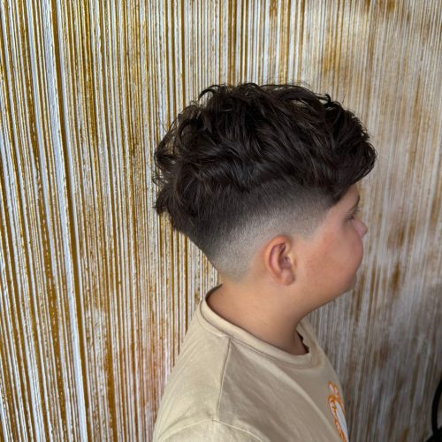 Imagine in profil a unui baiat cu par cret si voluminos deasupra, combinat cu un skin fade foarte curat pe laterale, cu trecere fina spre piele. Linia de contur este bine definita si precisa. Baiatul poarta un tricou bej si este fotografiat in fata peretelui decorativ auriu specific salonului Sia's Beauty din Lunca Cetatuii. Tunsoarea a fost realizata la frizerie copii Iasi, la Sia's Beauty.