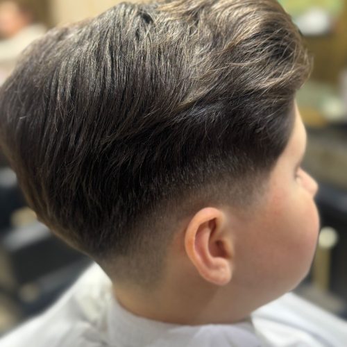 Imagine din spate si lateral a unui baiat mic cu o tunsoare tip pompadour, cu par lung deasupra dat spre spate cu volum si coafat ingrijit, combinat cu un degradeu curat pe laterale si ceafa. Linia de separatie dintre parul de deasupra si zona trasa la masina este precisa si bine definita. Baiatul poarta un halat alb de protectie. In fundal se observa spatiul luminos al salonului Sia's Beauty din Lunca Cetatuii, unde frizerie copii Iasi include si modele mai elaborate pentru cei mici.