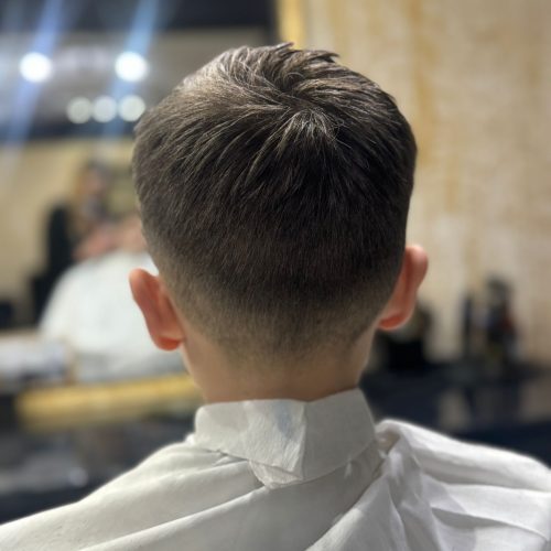 Imagine din spate a unui baiat cu o tunsoare ingrijita, cu par mai lung deasupra pieptanat si usor dat intr-o parte, si un degradeu low fade curat pe laterale si ceafa. Conturul cefei este bine definit si uniform. Baiatul poarta un halat alb de protectie cu servetel la gat. In fundal se observa oglinda cu rama aurie si luminile salonului Sia's Beauty din Lunca Cetatuii. Tunsoarea a fost realizata la frizerie copii Iasi, la Sia's Beauty.