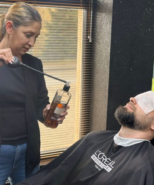 O specialista din echipa Sia's Beauty aplica un produs profesional L'Oreal Professionnel pe capul unui client barbat, folosind un aplicator dedicat. Clientul sta relaxat in scaunul de barbershop, cu un prosop de protectie pe frunte si un halat L'Oreal Professionnel. In fundal se observa fereastra salonului cu lumina naturala. Imaginea surprinde un moment din procesul de ingrijire profesionala la barbershop Iasi, la Sia's Beauty din Lunca Cetatuii.