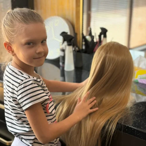 Anastasia, fiica patronilor salonului Sia's Beauty Center din Iasi, Lunca Cetatuii, privind zambitor spre camera in timp ce aranja parul unui cap de manechin. In fundal se observa interiorul salonului cu produse profesionale si oglinzile specifice unui spatiu de coafura.